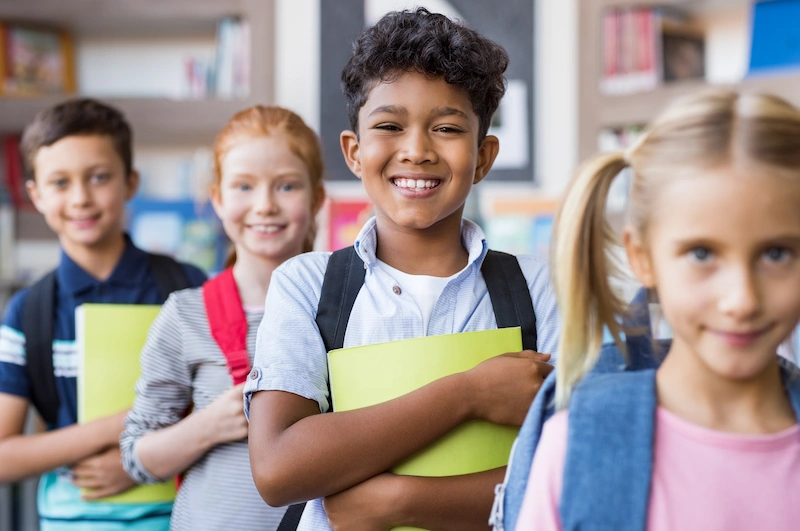 alunos em fila segurando o material escolar e sorrindo para a camera